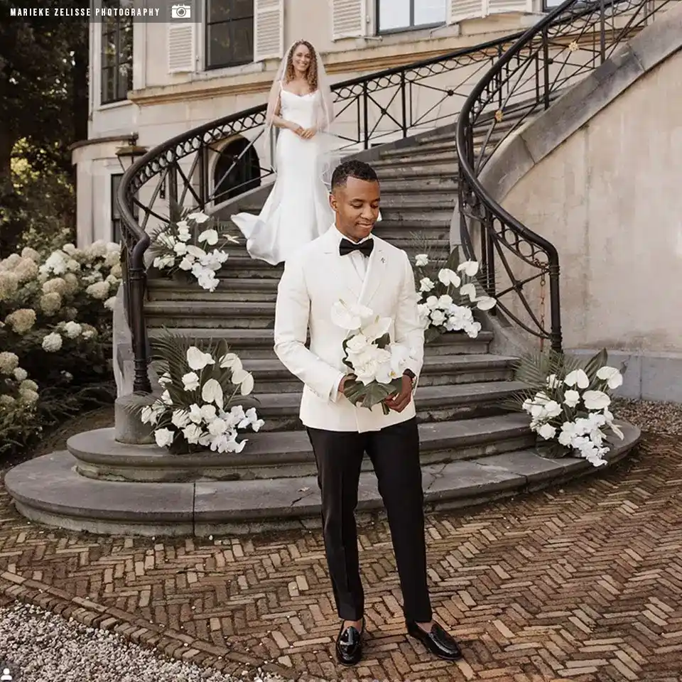 Rietbergh-klanten-vierkant-0001 Bruidegom in witte smoking van Rietbergh loopt voorop tijdens bruiloft, met bruid op trap van klassiek landhuis