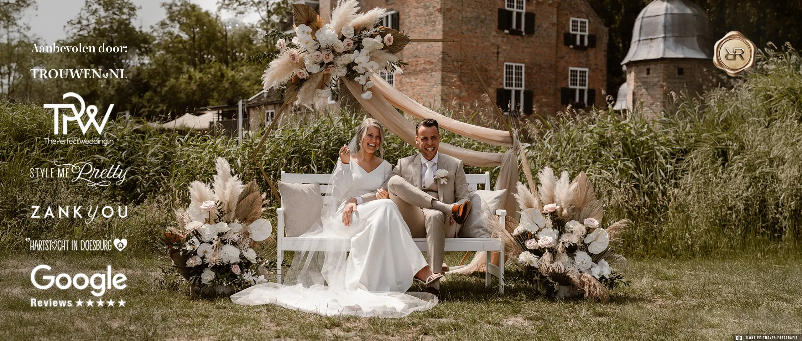 Een bruidspaar in een beige trouwpak en een witte jurk zittend op een bankje voor een kasteel.
